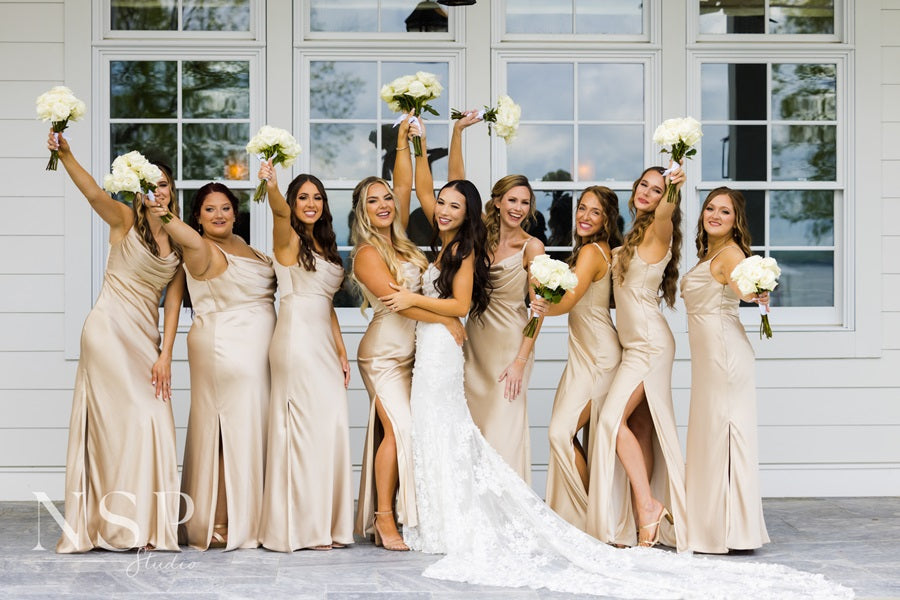 Bride and bridesmaids all holding their bouquets in the air. Bridal party is in shimmering beige satin dresses.