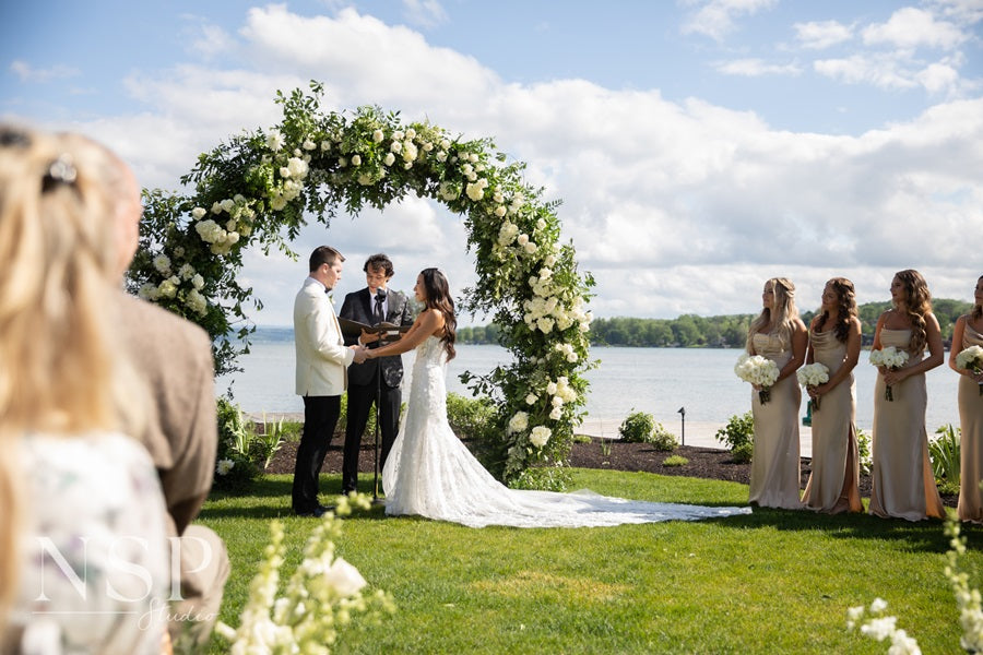 Bride and groom with the officiant under a large and lush floral archway full of whites and greens. Bridesmaids stand on the right holding their bouquets. The Canandaigua lake spans the background.