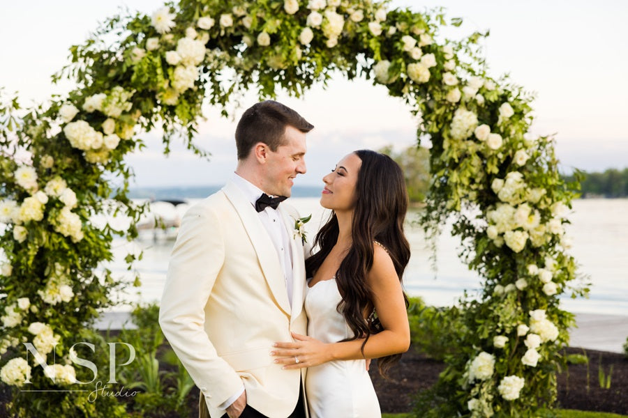 Bride and groom embrace in front of their lush floral arch in front of the lake at Lake House on Canandaigua.