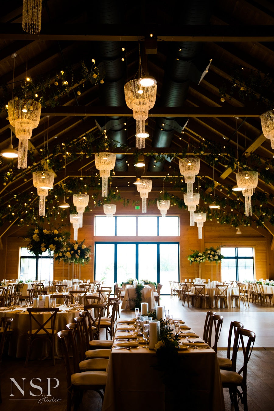 Room shot of the reception. Chandeliers hang from the ceiling with greenery and string lights wrapped around the rafters. Tables are set with greenery, florals, candles, and tableware.