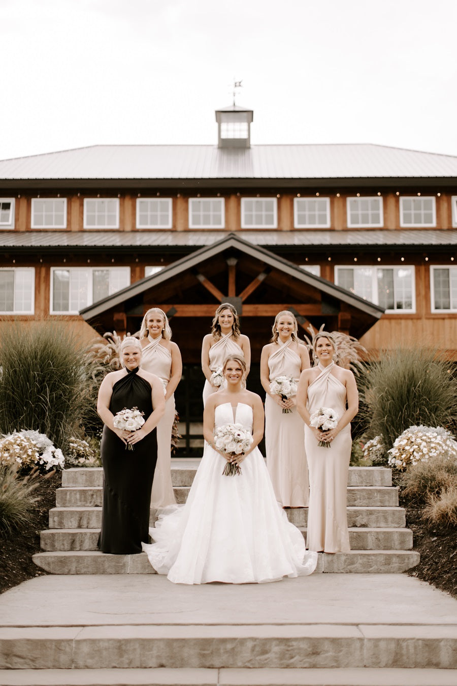 Bride and bridesmaids stand on steps in front of the Colloca Estate Winery. Bride is in white, maid of honor is in black, and the bridesmaids are in cream.