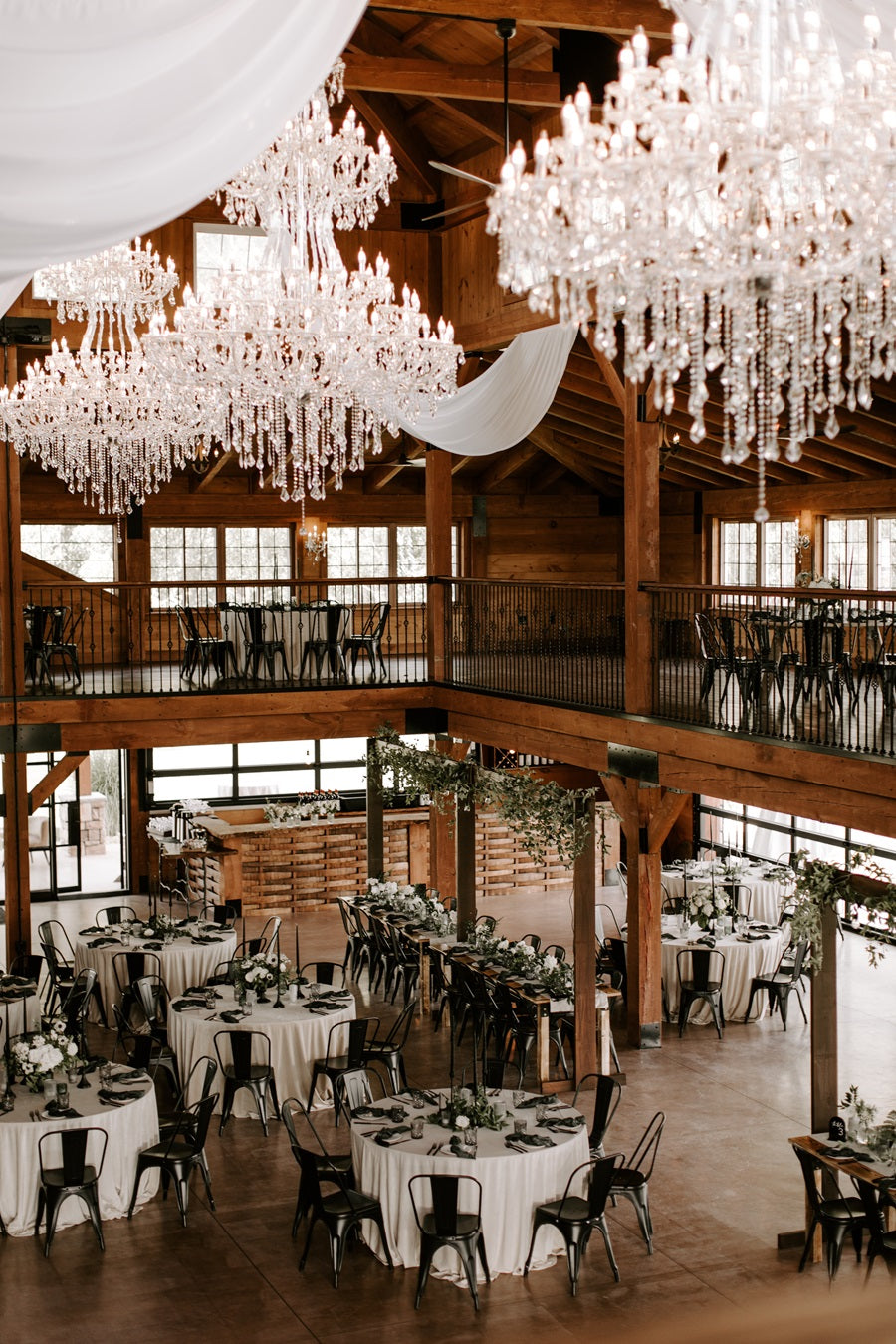 Top down view of the reception hall of the Colloca Estate Winery. Three grand chandeliers hang between floors, round tables set in the white, green, and black color palette line the floor.