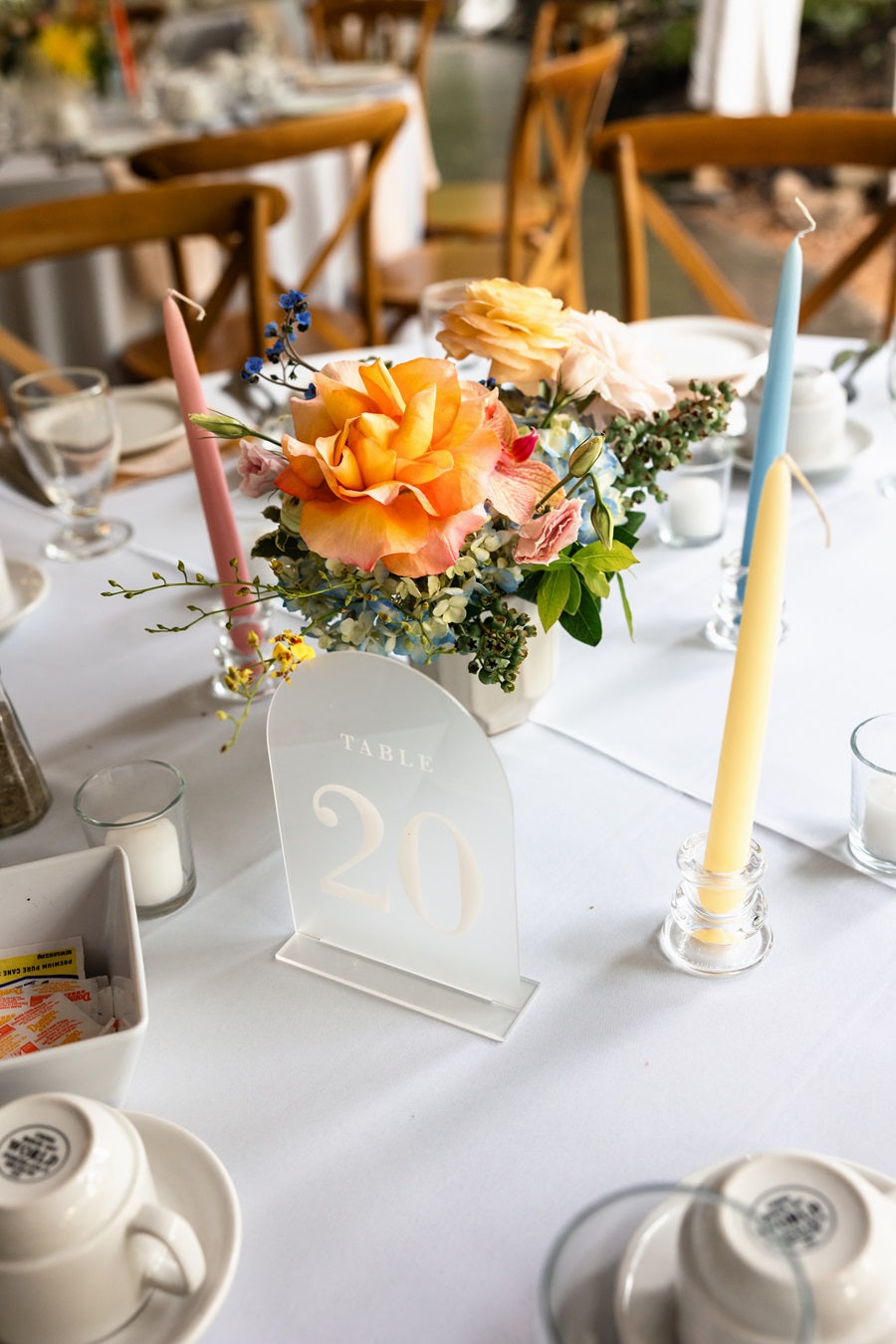 Close up on a table setting, highlighting a colorful centerpiece with roses, hydrangea, greenery, and more. Accented with pink, yellow, and blue taper candles.