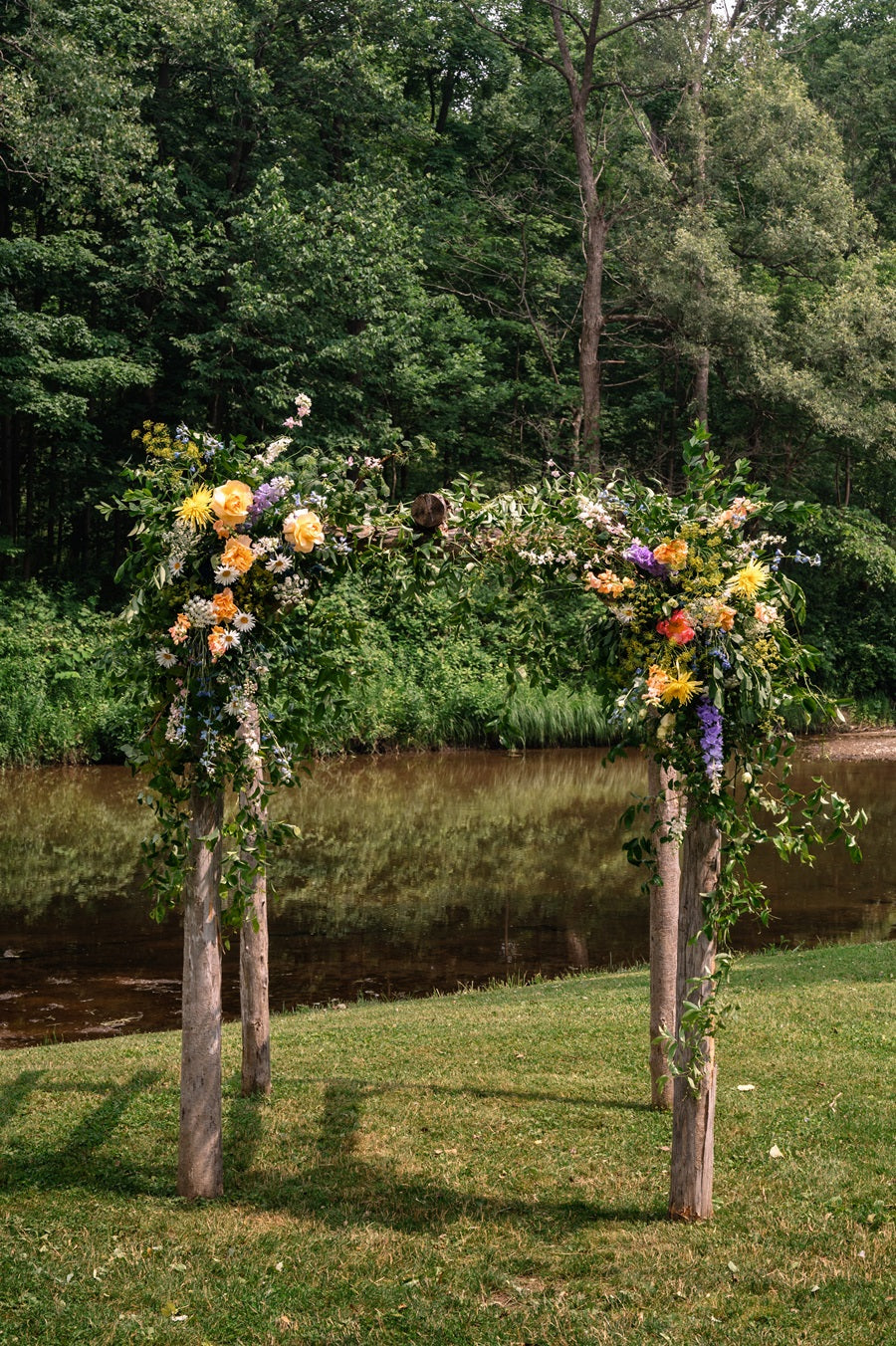 An arch stands along a forest and a river. It has a natural feel, full of greenery and florals. The florals shown are yellow, orange, purple, and pink, with blue accents.