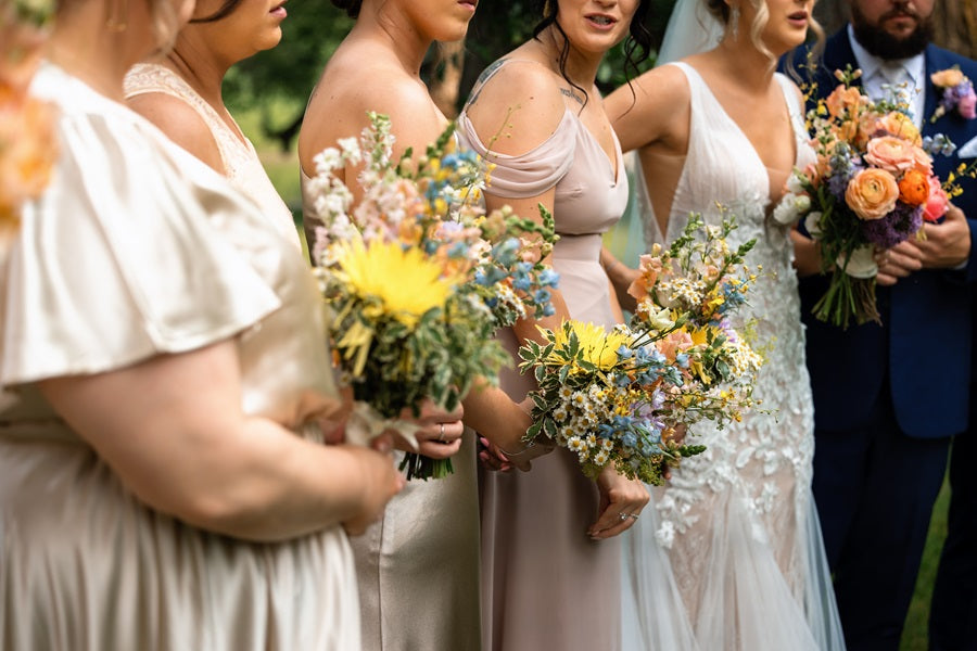 Bridal party stands in light pink and silver, holding small bouquets of yellow, green, blue, and pink accents. Bride and groom are in the background with matching rainbow colored florals.