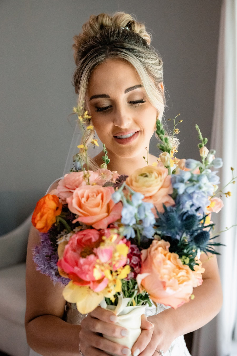 Bride holds up colorful bridal bouquet full of purple, pink, orange, blue, and yellow. Some of the florals pictured are roses, delphinium, and blue thistle.