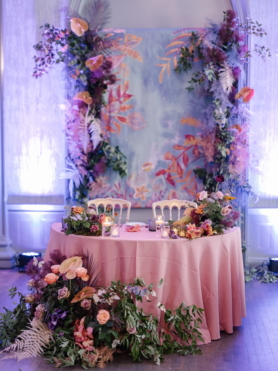 The sweetheart table with a pink table cloth, florals framing the bottom and a floral backdrop. The table gives off a whimsical fairytale garden aesthetic. Purple lights are lit to add to the atmosphere.