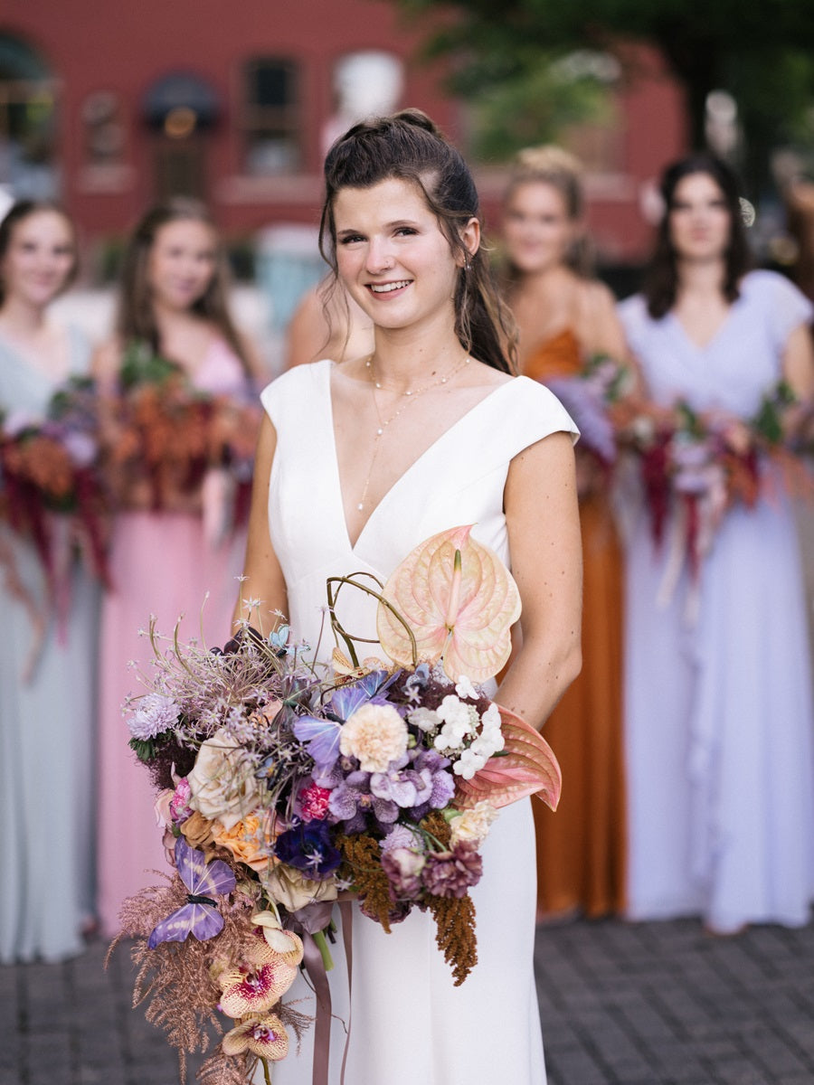 Stand alone shot of the bride in front of her bridal party. She is in a white dress with a cascading bouquet of purple, yellow, brown, and orange.