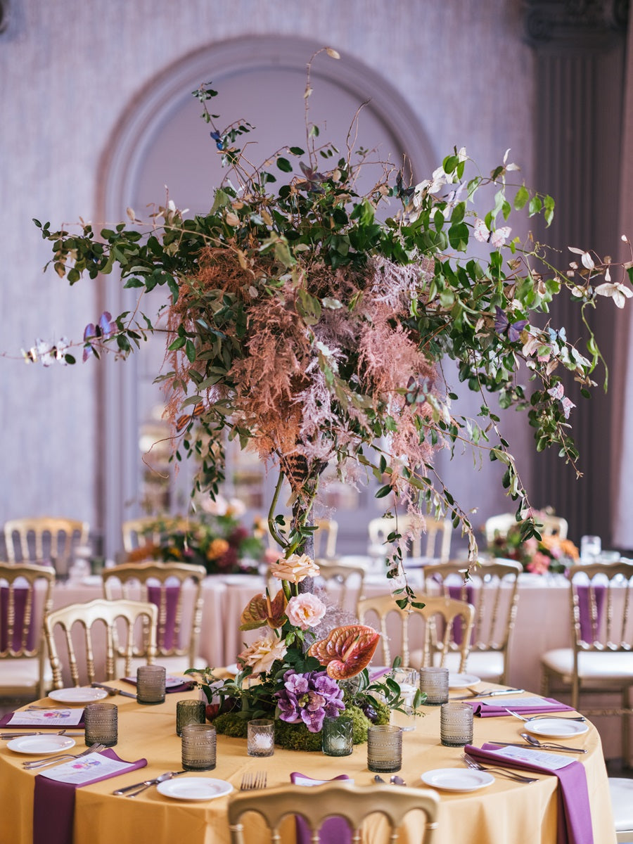 Close up on the tall centerpiece. It is on a stand lifting above the table. The arrangement is lush with greenery and accent colors such as pink and purple. It is in a whimsical fairytale garden style.