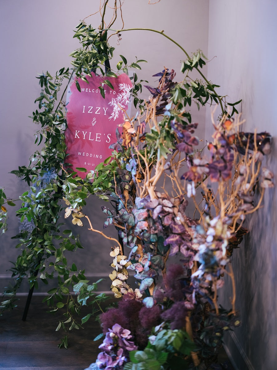 A greenery and butterfly focused welcome sign with branching. Whimsical and natural feel. Pink sign with green, blue, purple, and yellow butterflies.