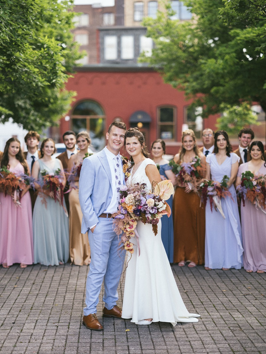 Shot of the bride and groom in front of the bridal party. Bridal party is in a variety of colored dresses within the wedding color palette (pink, orange, green, yellow, blue). Bride is holding a cascading bridal bouquet