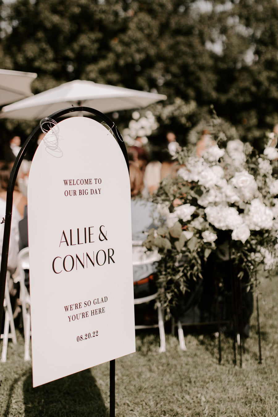 Welcome sign "welcome to our big day" "Allie & Connor" "We are so glad you are here, 8.20.22" whites and greens floral arrangement placed at the end of the aisle in the background.