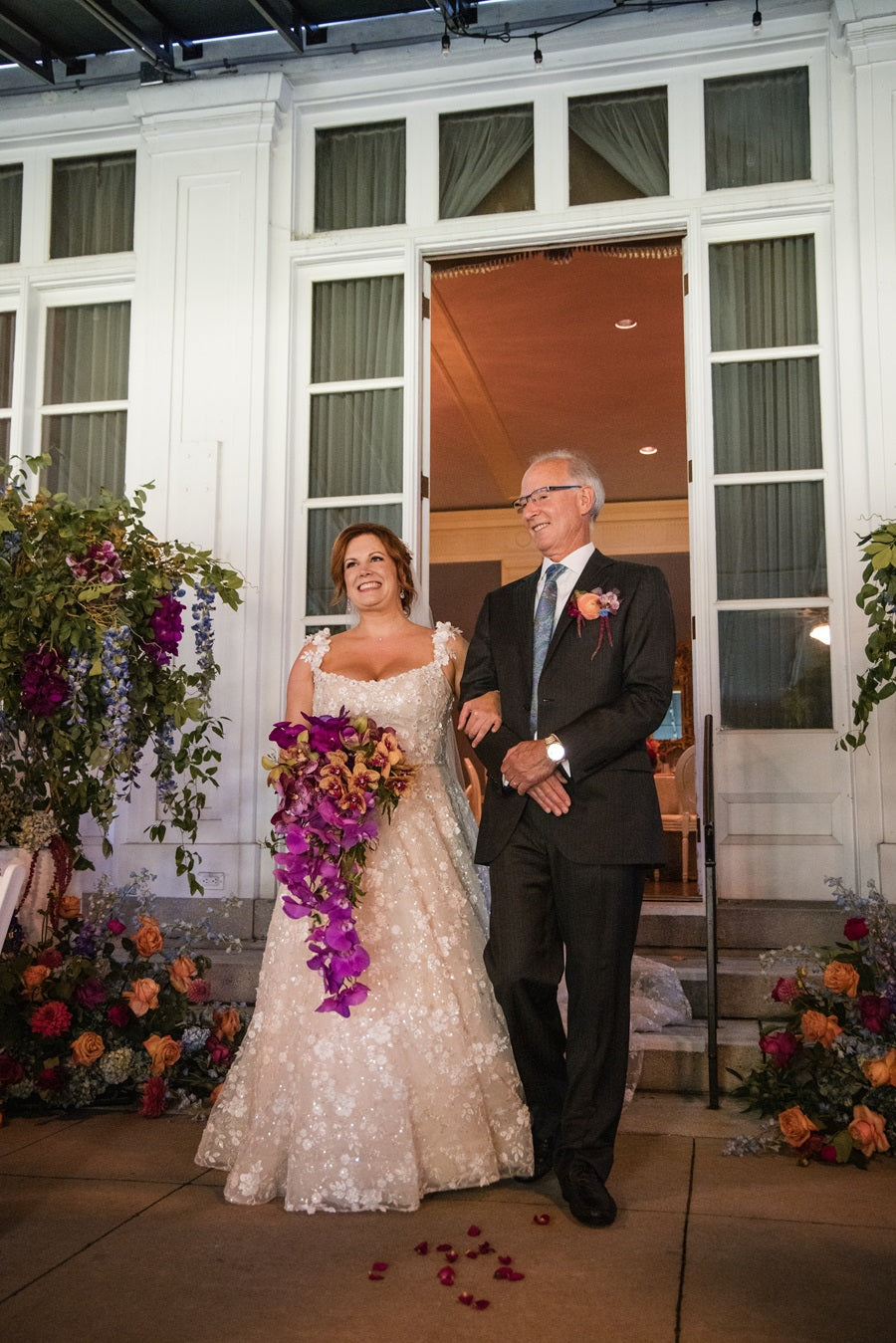 Bride and father walk down the aisle. Bride holds a bouquet full of orchids. The aisle entrance is lined with a floral arrangement and tree with floral accents. Some flowers shown are orchids, roses, hydrangea, and delphinium. Jewel toned color theme.