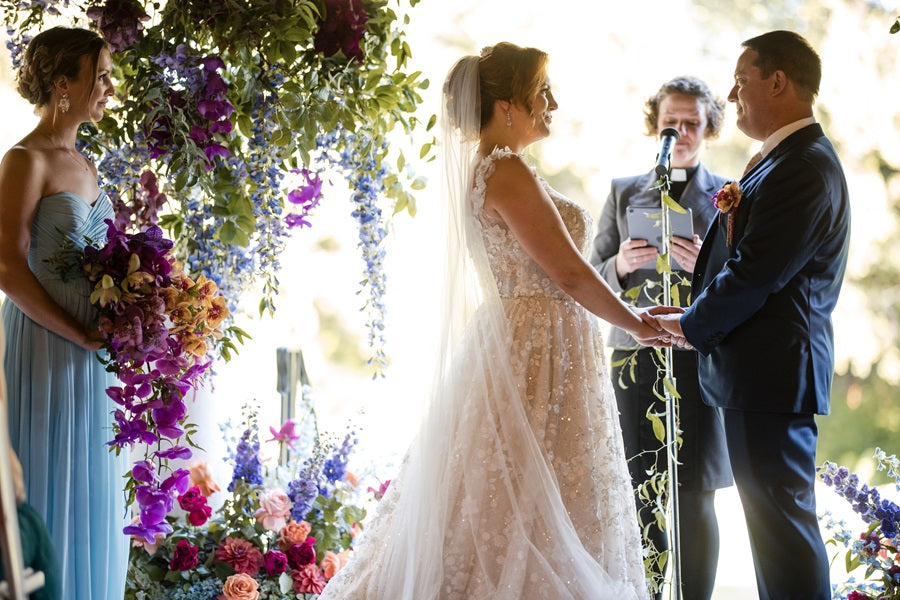 Bride and groom stand together with a large floral arch pieces in a jewel toned color palette. MOH holds the bridal bouquet of orchids of to the left.