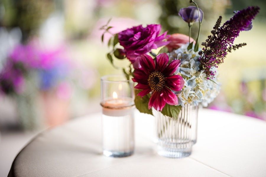 Close up on a small centerpiece on a cocktail table. The florals are pink, blue, and purple. Some florals pictured are roses and hydrangea.