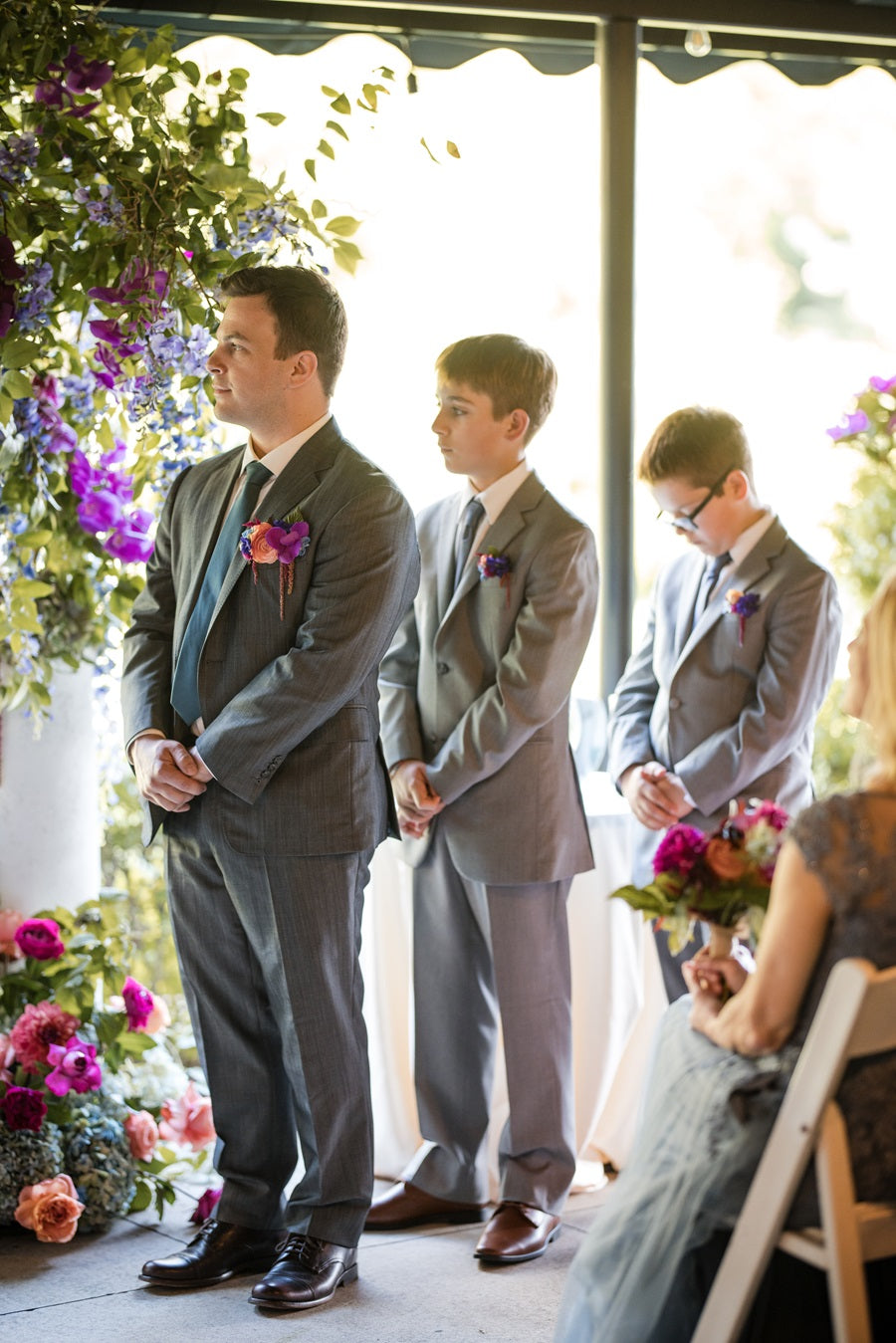 Groomsmen stand to the right of the groom, hands fold in front. They all wear matching pocket squares of pink, pink, purple, and blue, with green accents.