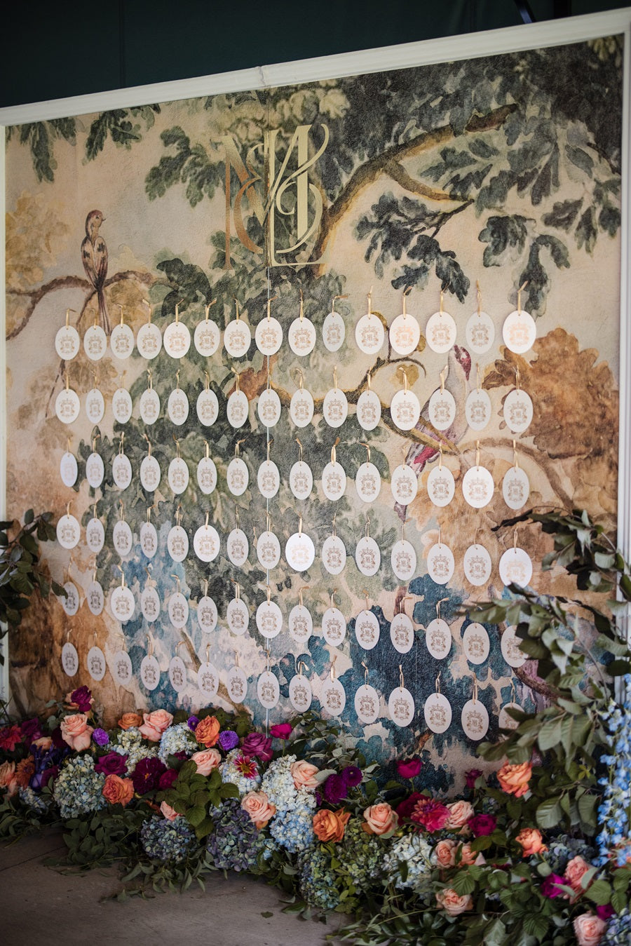 The guest table seating wall is covered in a watercolor floral pattern with fresh floral lining the floor in front. The colors match the jewel tone theme of the wedding and feature roses, hydrangea, orchids, delphinium, greenery, and more.