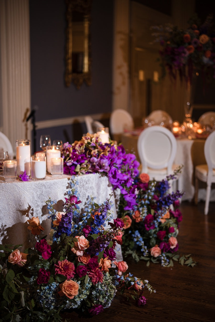 The sweetheart table has lush florals on the top draping to the floor with large floral arrangements lining the floor in front. The focal florals are pink orchids, and hydrangea/roses/delphinium in pink/blue/purple/peach.