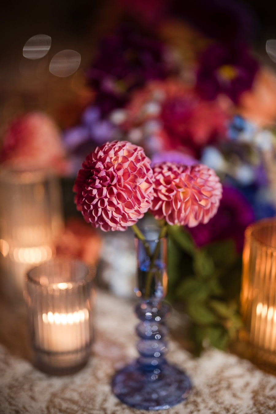 Close up on a purple glass bud vase with two pink dahlias. A larger floral centerpiece and candles can be seen in the background.