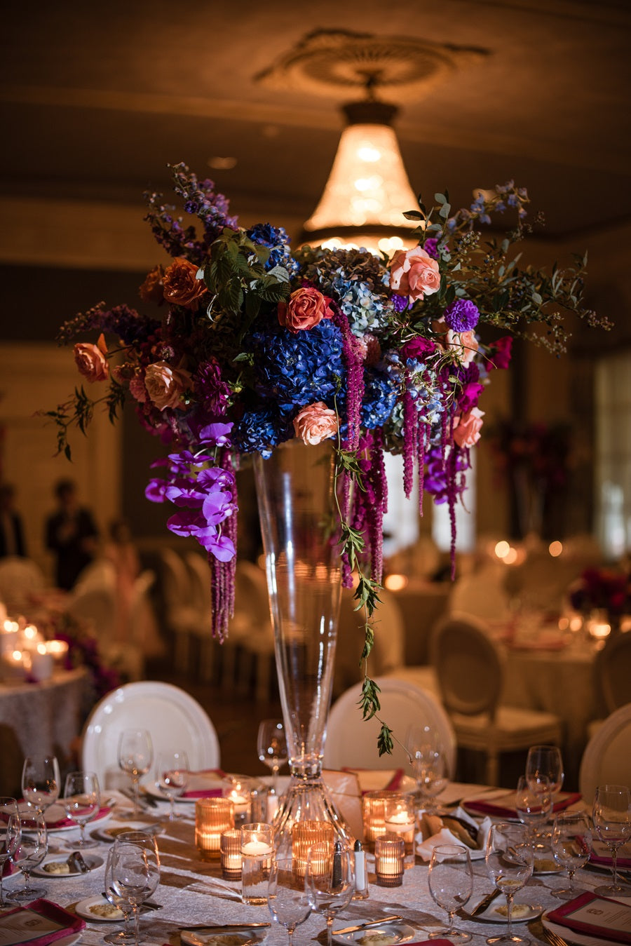 A large raised centerpiece. Lifted on a glass stand. Florals are jeweltoned, featuring hydrangea, roses, orchids, delphinium, and greenery.