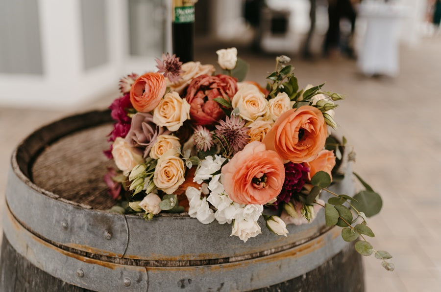 A floral arrangement on a barrel. The arrangement is lush with blooms such as peonies, roses, and zinnia.