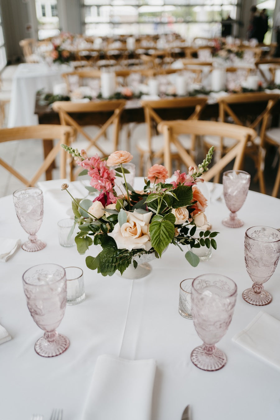 A floral centerpiece with greenery and florals. Some florals shown are roses and snapdragons. Placed on a round table with set tableware.