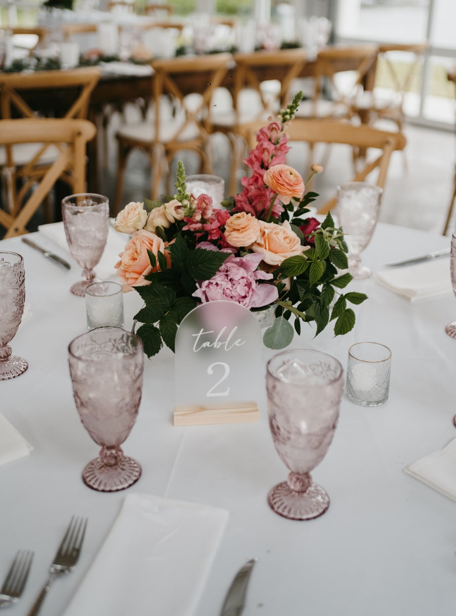 A floral centerpiece on a round table, colors pink, peach, and green. Table is labeled #2 and set with tableware.