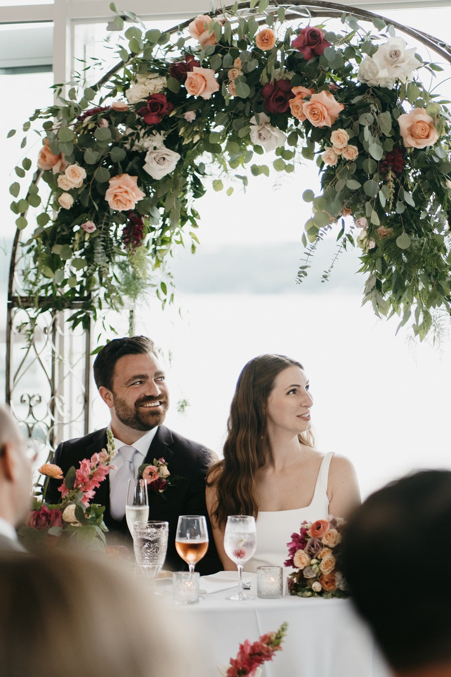 Bride and groom sitting at the sweetheart table during the reception. The Ceremony floral arch frames their background (having been brought inside the venue).