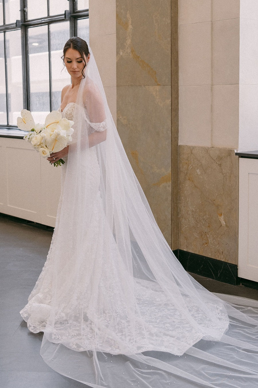 Bride stands alone, looking down to her side. She holds a bridal bouquet full of roses and calathea.