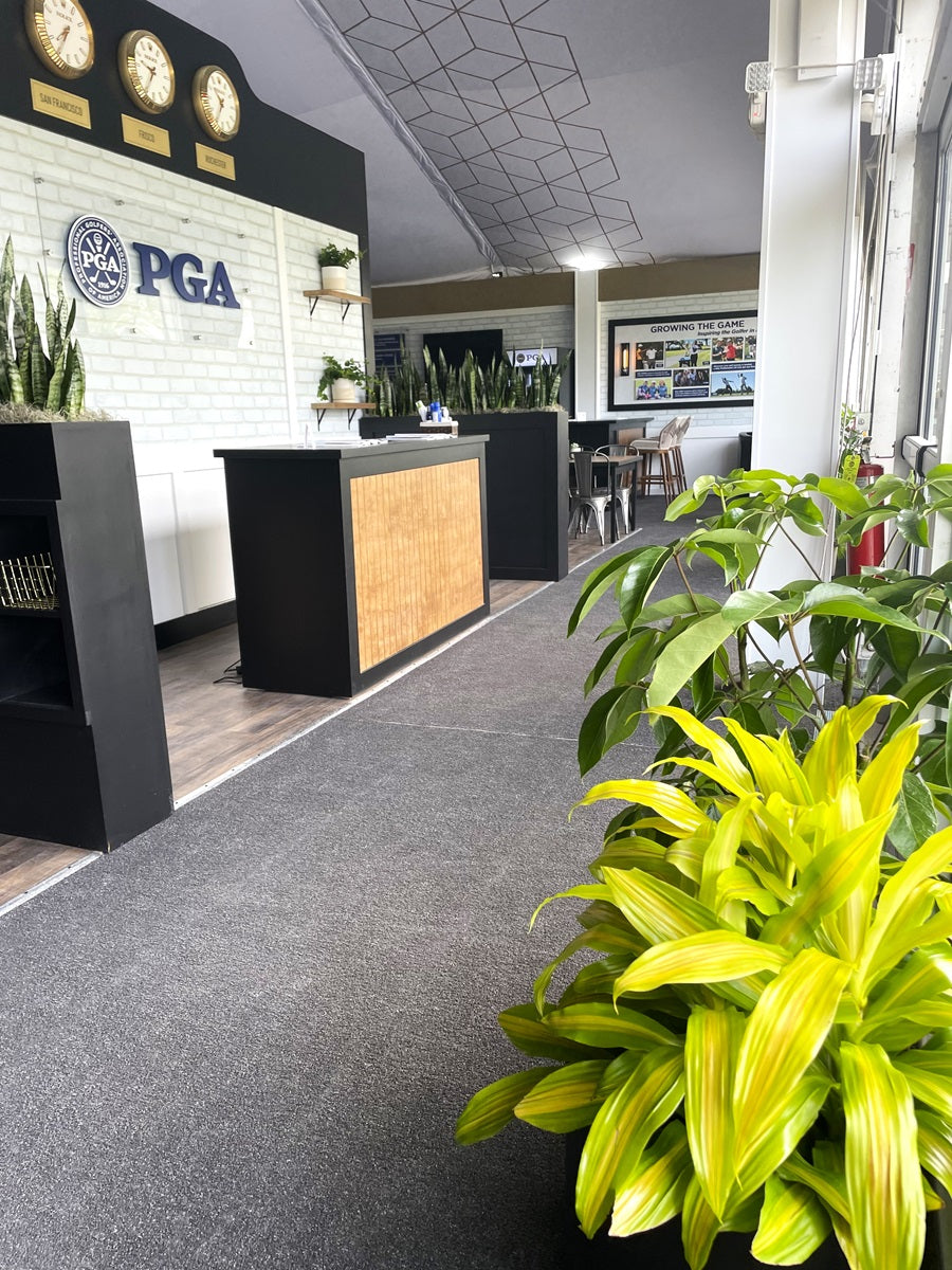 Shot of PGA front desk at Oak Hill Country Club. Large plants are placed next to the entrance, plants scaping with moss fill planters on either side of the desk. The PGA logo displayed in the back with clocks showing different time zones.