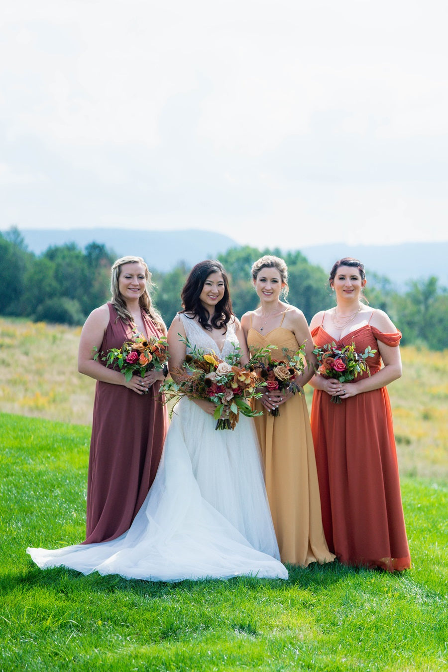 Bride and bridesmaids standing together holding their bouquets. Bride is in white, bridesmaids are in various colors. From left to right, burgundy red, yellow, and an orange/red.