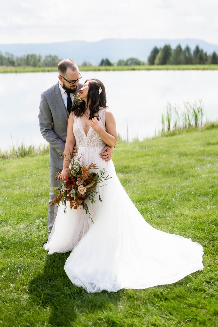 Bride and groom embrace in front of a lake at the Wrens Roost Barn. Bride holds a bouquet made in fall colors.