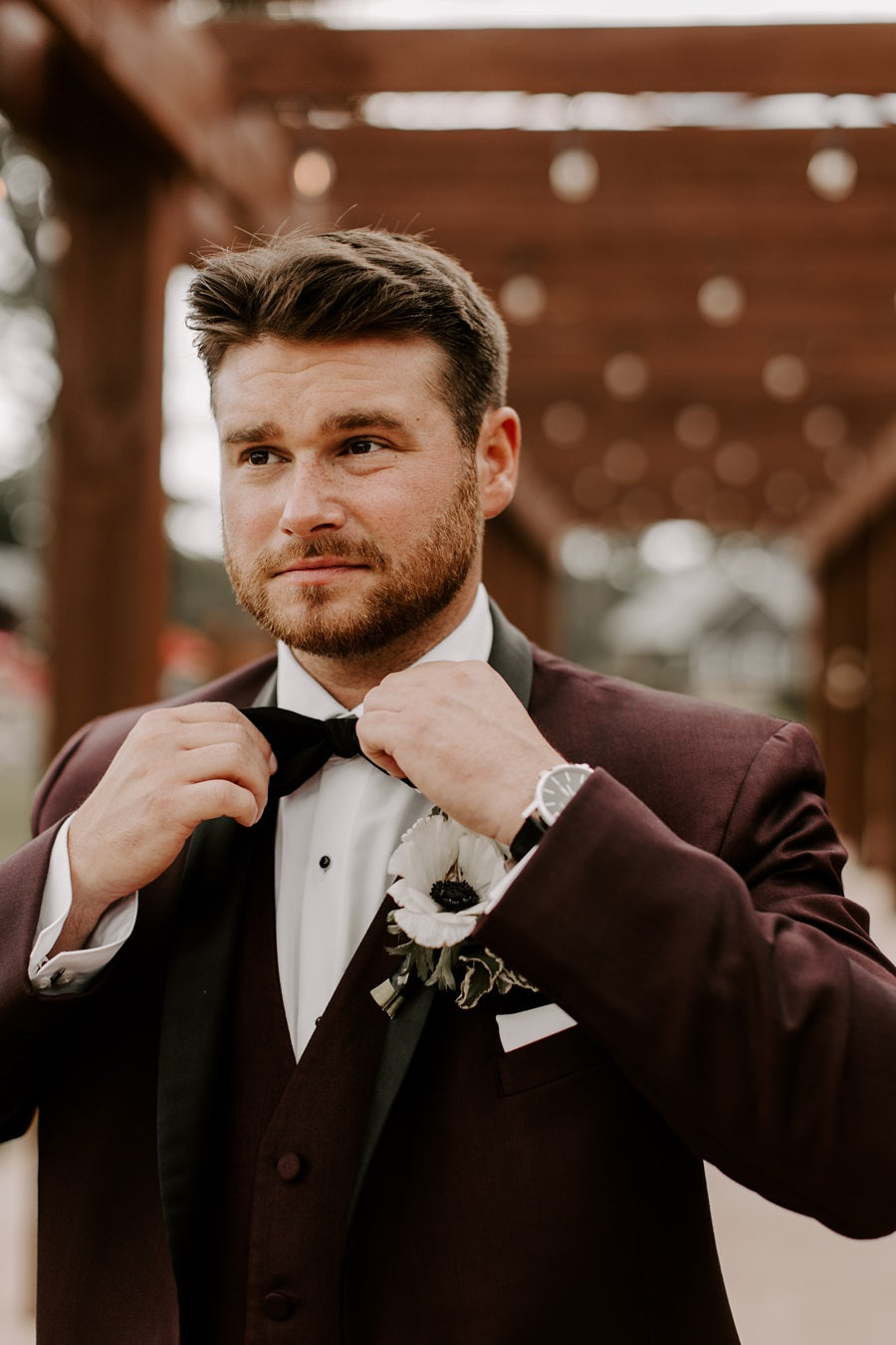 Groom adjusting his bow tie. He is in a dark burgundy suit with a white/black floral boutonniere.