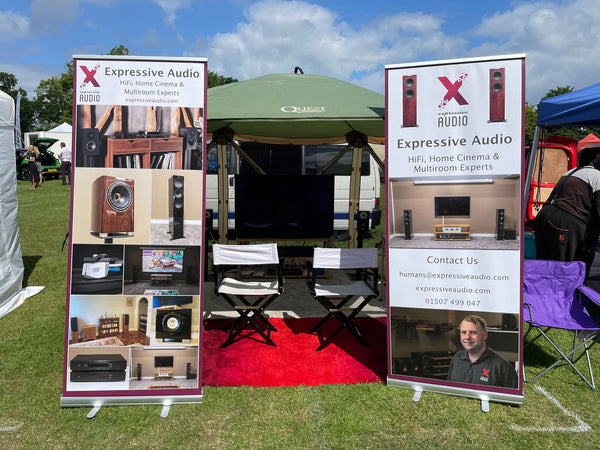 A gazebo tent outdoors with a HiFi system set up inside it, with two chairs just inside the entrance. Outside the tent are two banners advertising Expressive Audio.