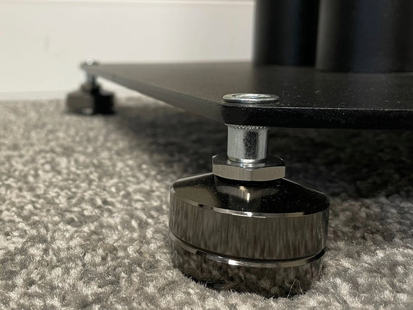 A close up photo of an IsoAcoustics GAIA I threaded isolation foot under the corner of a speaker stand, with another one visible but out of focus in the background to the left.