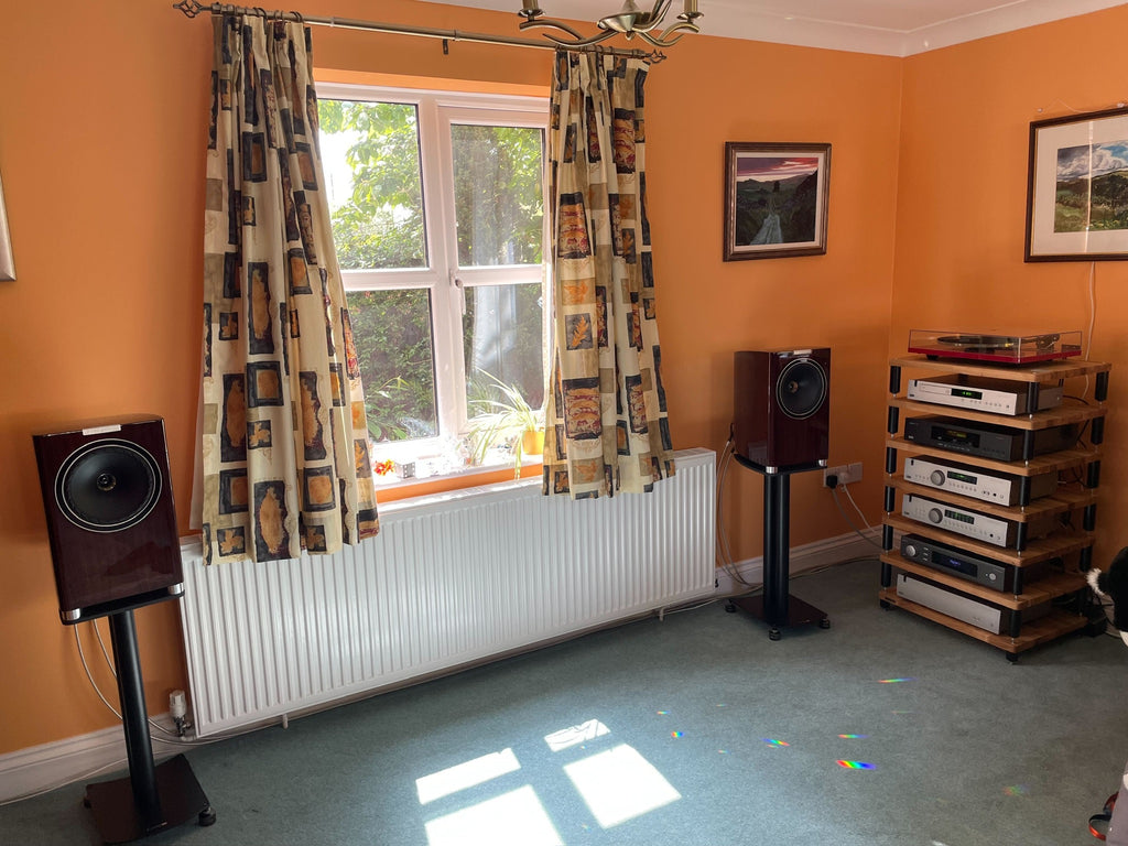 A photo of a HiFi system in a room of a house which has orange painted walls and green carpet. At the right hand side there is a HiFi rack with HiFi components stacked on it, and there are loudspeakers either side of a window which is in the middle of the photo.