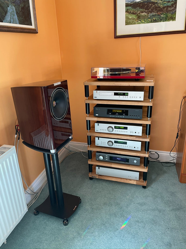 A photo of a HiFi rack with HiFi components stacked on it, and a loudspeaker to the left of it. They are in the corner of a room, and the walls behind are painted orange and the carpet is green.