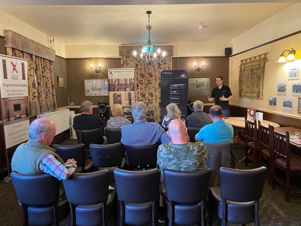 A photo of a room in a pub with three rows of chairs set up and people sitting on them facing the front of the room, where there is a HiFi system set up. There are two banners either side of the HiFi system, one advertising Expressive Audio and the other advertising Cyrus Audio. At the right hand side is a person with short hair, wearing a black polo top, speaking to the people who are sitting down.