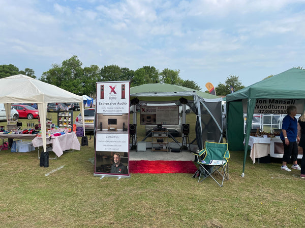 A photo of a green gazebo tent with a HiFi system set up inside it. Outside the tent entrance there are two banners advertising Expressive Audio. On either side of the tent are other tents.