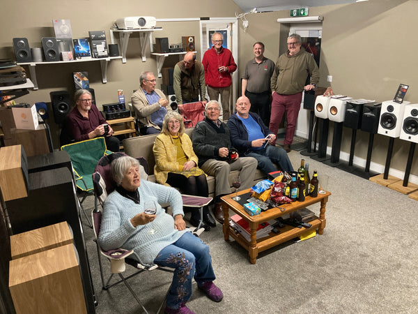 A group of people sitting in Expressive Audio's HiFi showroom, with a table of drinks and snacks in front of them and HiFi equipment along the walls around them.