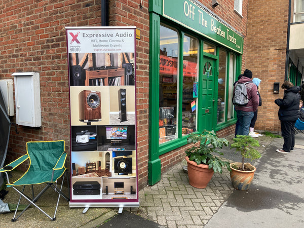 A photo from the side of the green shop front of Off The Beaten Tracks, with a sign advertising Expressive Audio to the left and a green chair in the bottom left corner.