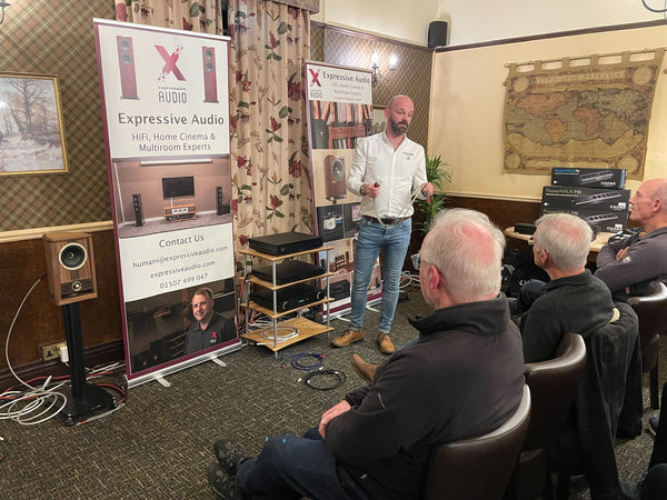 A photo of a person in a white shirt and pale blue jeans standing at the front of a room talking to a group of people sitting down. Behind the person at the front is a HiFi system set up on a wooden rack, with banners advertising Expressive Audio on either side.