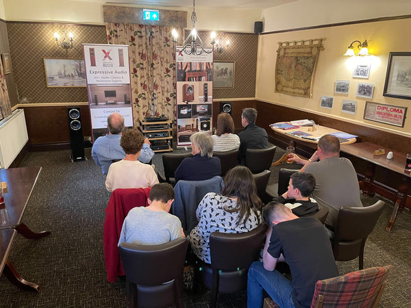 A group of people sitting in a room at a pub listening to music on a HiFi system set up at the front of the room. Either side of the HiFi are two banners advertising Expressive Audio.