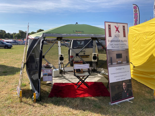 A photo of a green gazebo tent in a field with a HiFi system set up inside it, and banners advertising Expressive Audio outside it.