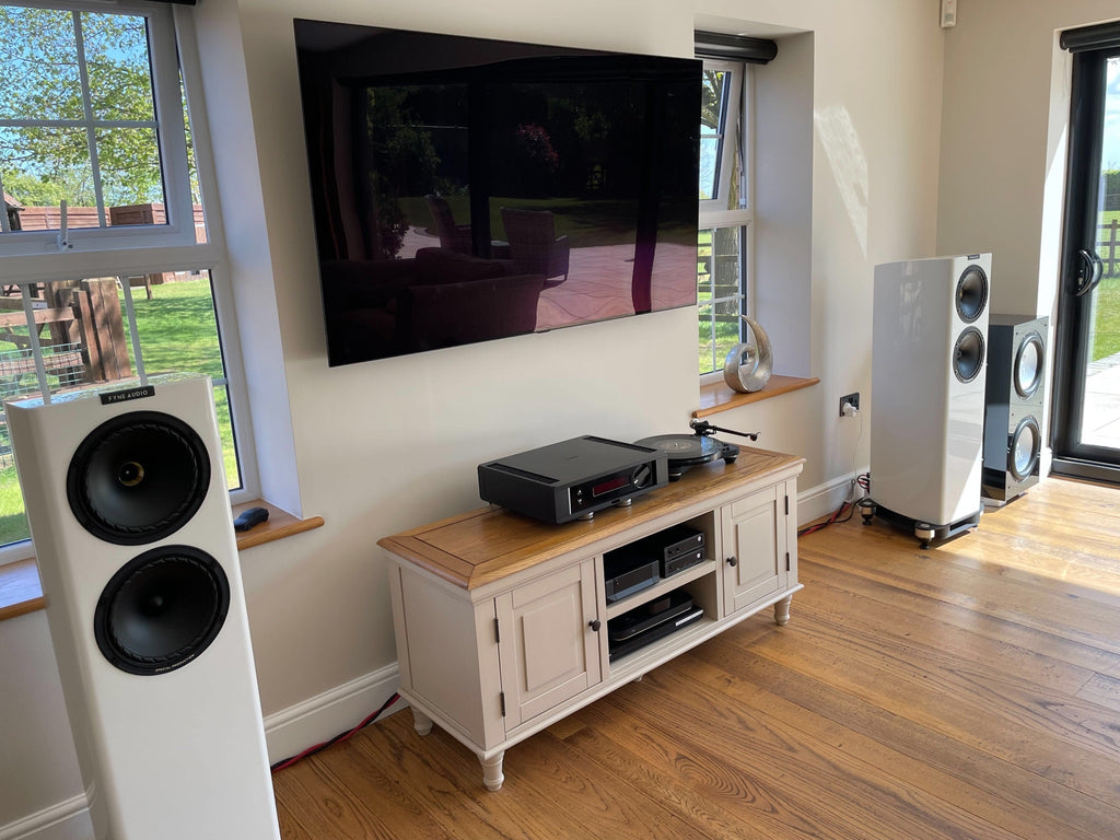 A photo of a HiFi system with a wall-mounted TV, under which is a white storage unit with wooden top and HiFi components on it, and white loudspeakers on either side.
