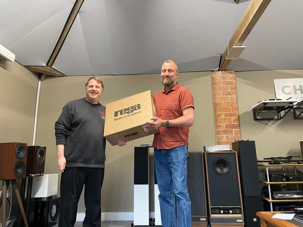 A photo taken inside a HiFi showroom, with a person on the left wearing a grey Expressive Audio branded sweatshirt, and a person on the right wearing an orange shirt and blue jeans. They are both smiling and between them holding a cardboard box with the brand name "Rega" on it.