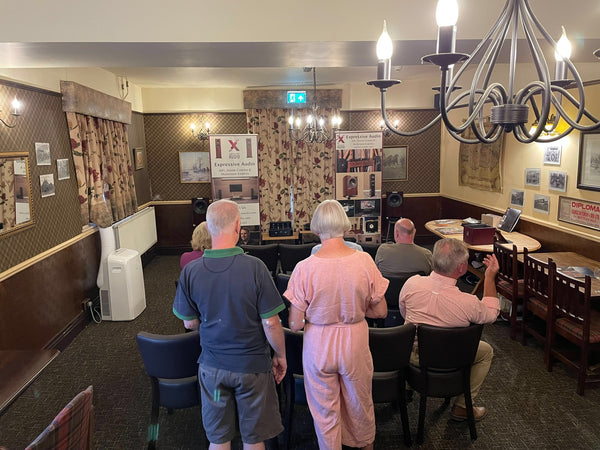 A group of people sitting and standing in a room at a pub listening to music on a HiFi system which is set up at the front of the room. There are two banners advertising Expressive Audio next to the HiFi.