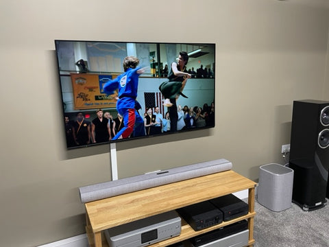 A photo of a TV screen mounted on a wall, with the Harman Kardon Citation MB1100 Soundbar on a wooden unit below it and the Harman Kardon Citation Sub S subwoofer on the floor to the right.