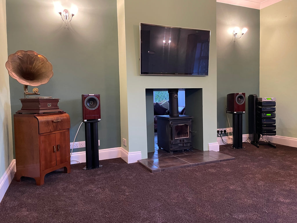 A photo of a HiFi system set up in a room in a house, with a TV mounted on the wall in the middle above an alcove wood burning stove, with loudspeakers on either side, a gramophone on a stand at the left, and a stack of HiFi components on a rack at the right.