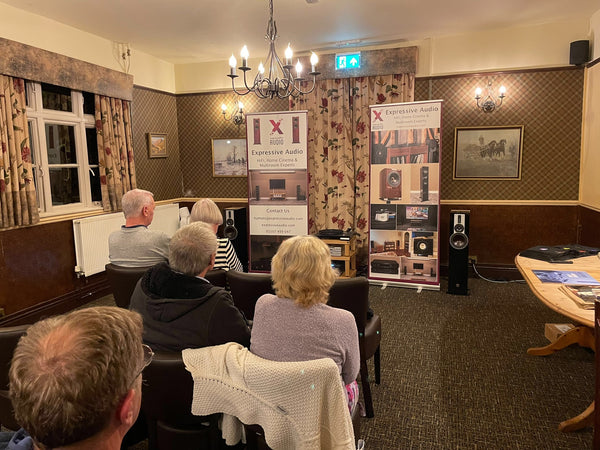 A group of people sitting in a room at a pub, listening to music on a HiFi system which is set up at the front of the room. Next to the HiFi are two banners advertising Expressive Audio.